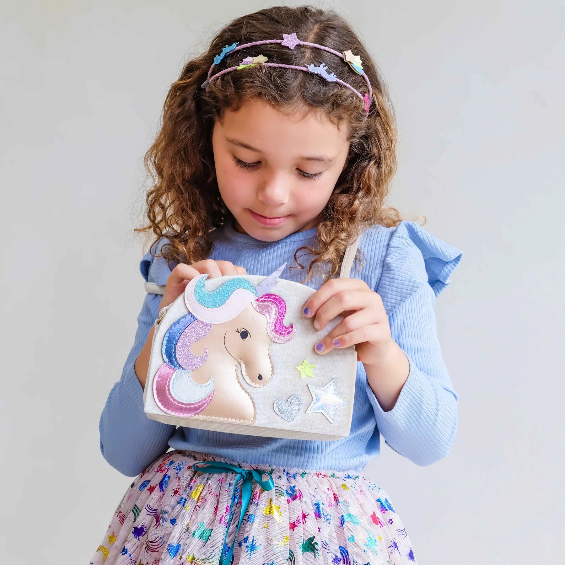 A young girl with curly brown hair wearing a pastel unicorn headband and a blue ribbed top holds a small white shoulder bag decorated with a colorful embroidered unicorn and glittery star details.