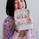 A smiling young girl with short black hair holds open a small beige jewelry organizer box. The box contains several colorful hair clips, and the girl is wearing a pink long-sleeve top with a shiny multicolor star-patterned overlay.