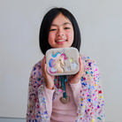 A smiling young girl with short black hair holds up a small rectangular pouch or case decorated with a colorful unicorn applique, star details, and a heart-shaped charm. She is wearing a pink top and a sheer pastel cardigan covered in small multicolored butterfly and rainbow patterns, against a plain light gray background.