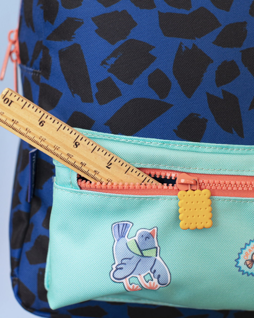 Blue backpack with black pattern and turquoise pouch featuring a bird design, held open by a ruler.