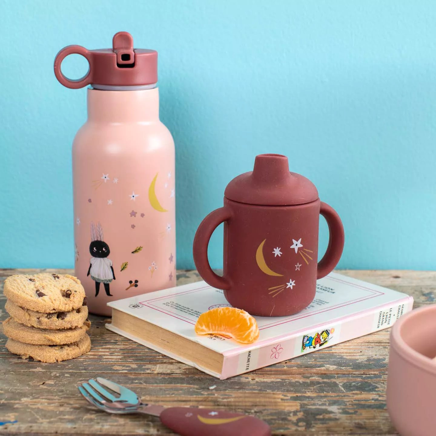 Moulin Roty Pink water bottle and red sippy cup on a wooden table with cookies and an orange.