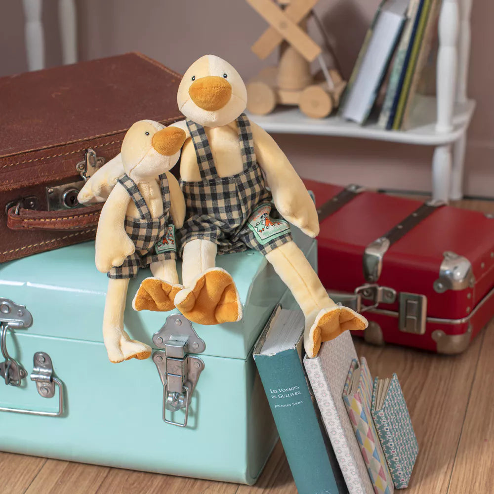 Two toy ducks in checkered overalls sitting on a suitcase with vintage suitcases and books in the background.