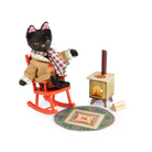 Toy black cat in a sweater sitting on a rocking chair next to a small stove with a fire.