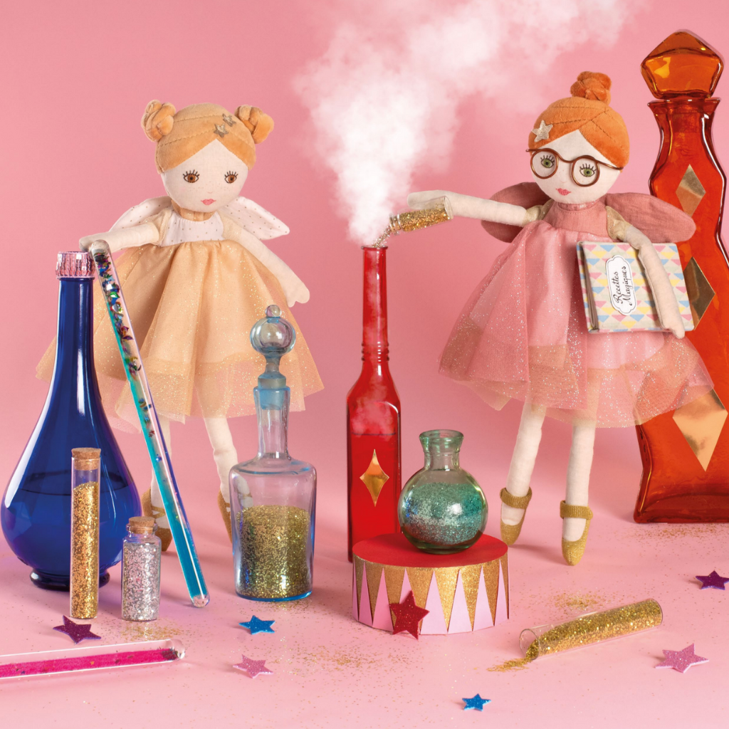 Two moulin roty fairy dolls with colorful bottles and props on a pink background