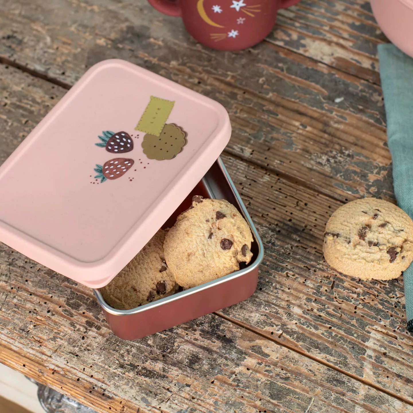 Small metal lunchbox for children with pink lid containing cookies on a wooden surface