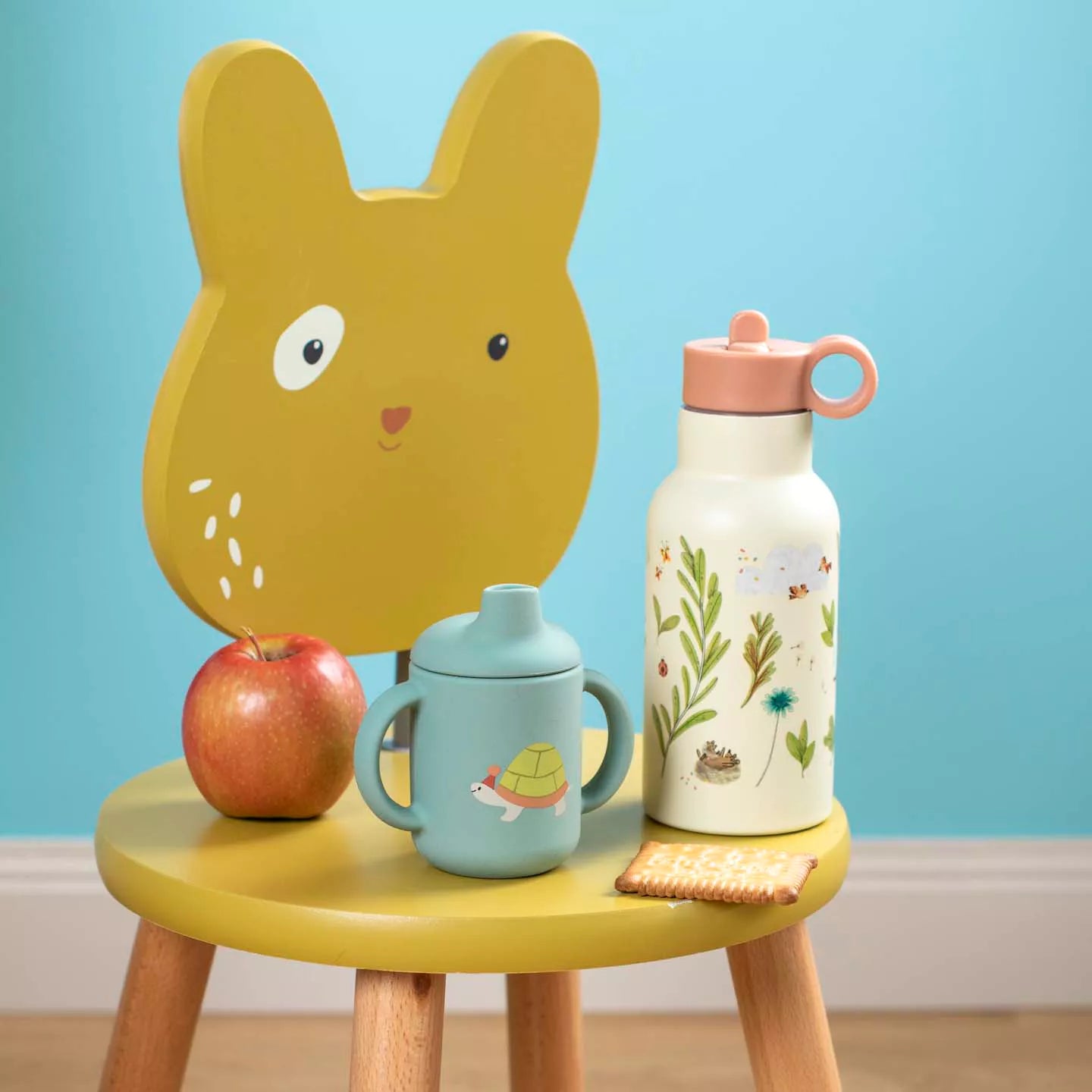 Yellow children's chair with a face, apple, mug, and water bottle on a blue background