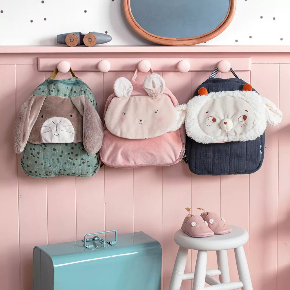 Three animal-themed childrens backpacks hanging on a pink wall with a teal box and white stool below.