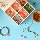 A colorful bead craft kit displayed in an open wooden multi-compartment box, surrounded by finished beaded jewelry and loose beads on a light blue background.