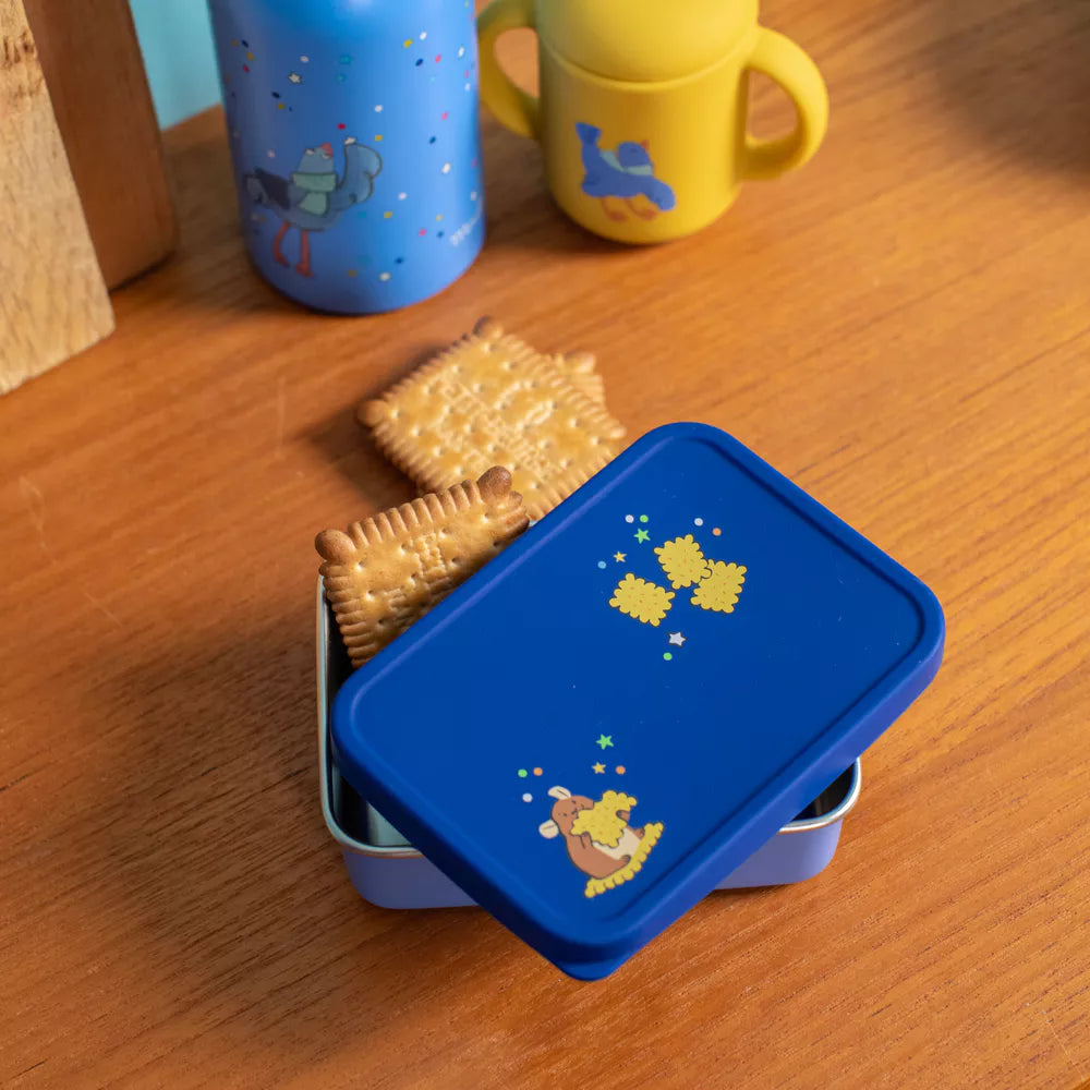 Blue lunch box with Moulin roty puce and pilou design containing crackers on a wooden surface with colorful cups in the background.