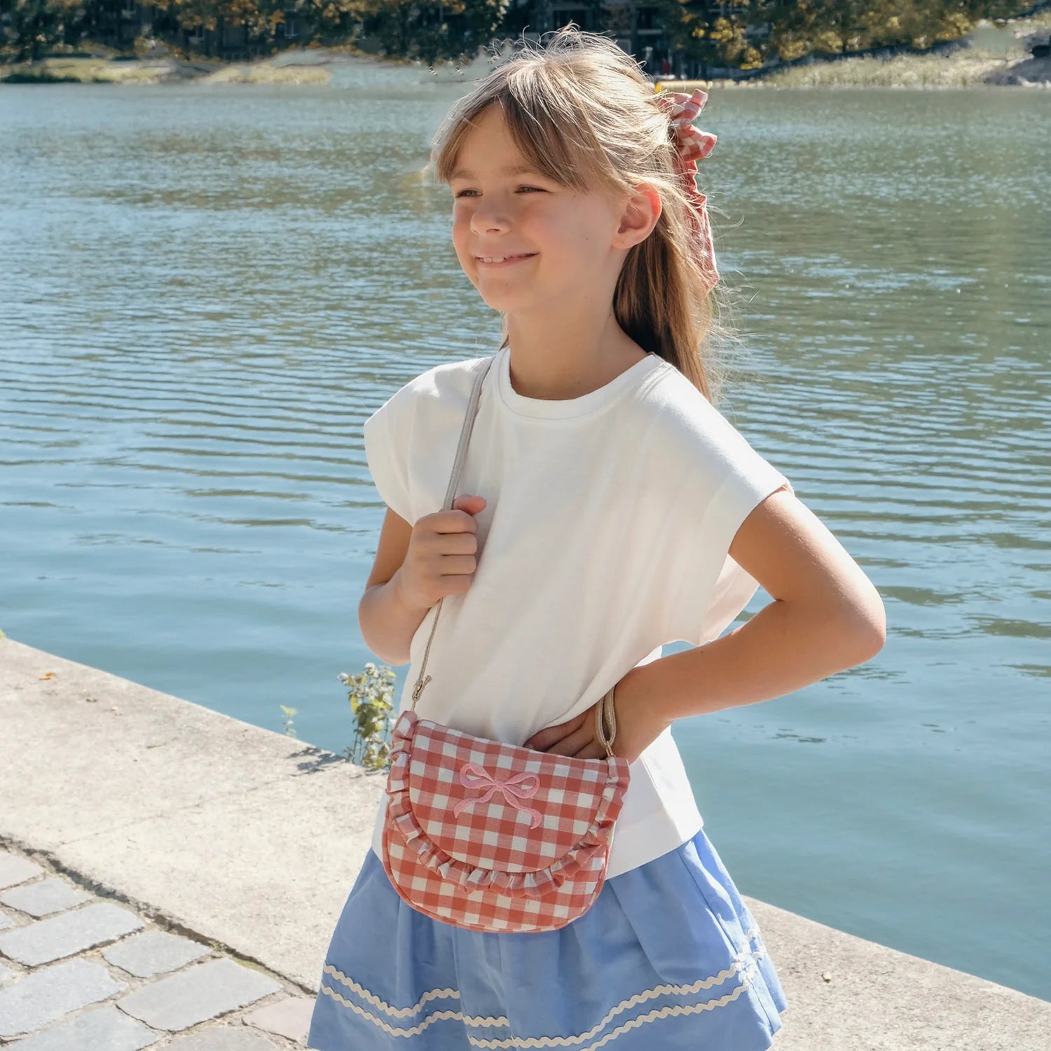 Young girl standing by a body of water wearing a white shirt and blue skirt with a red checkered bag.