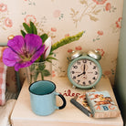 A styled tabletop scene with a light blue enamel mug, a mint green vintage twin-bell alarm clock, a small book, a wooden block, and a glass vase holding a bright purple flower and green stems, set against floral wallpaper.