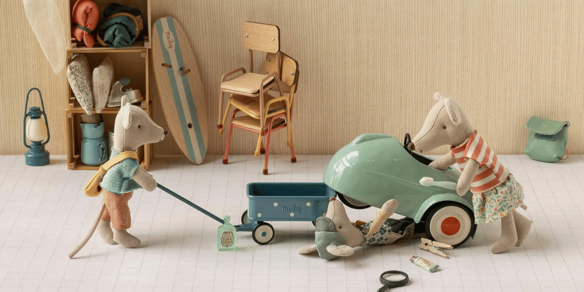 A cozy indoor play scene with plush animal characters: one pushes a blue wagon, one drives a mint green toy car, and another lies nearby on a light wood floor. The room has a striped wallpaper background, wooden shelves with beach-themed items, and a surfboard leaning against the wall.