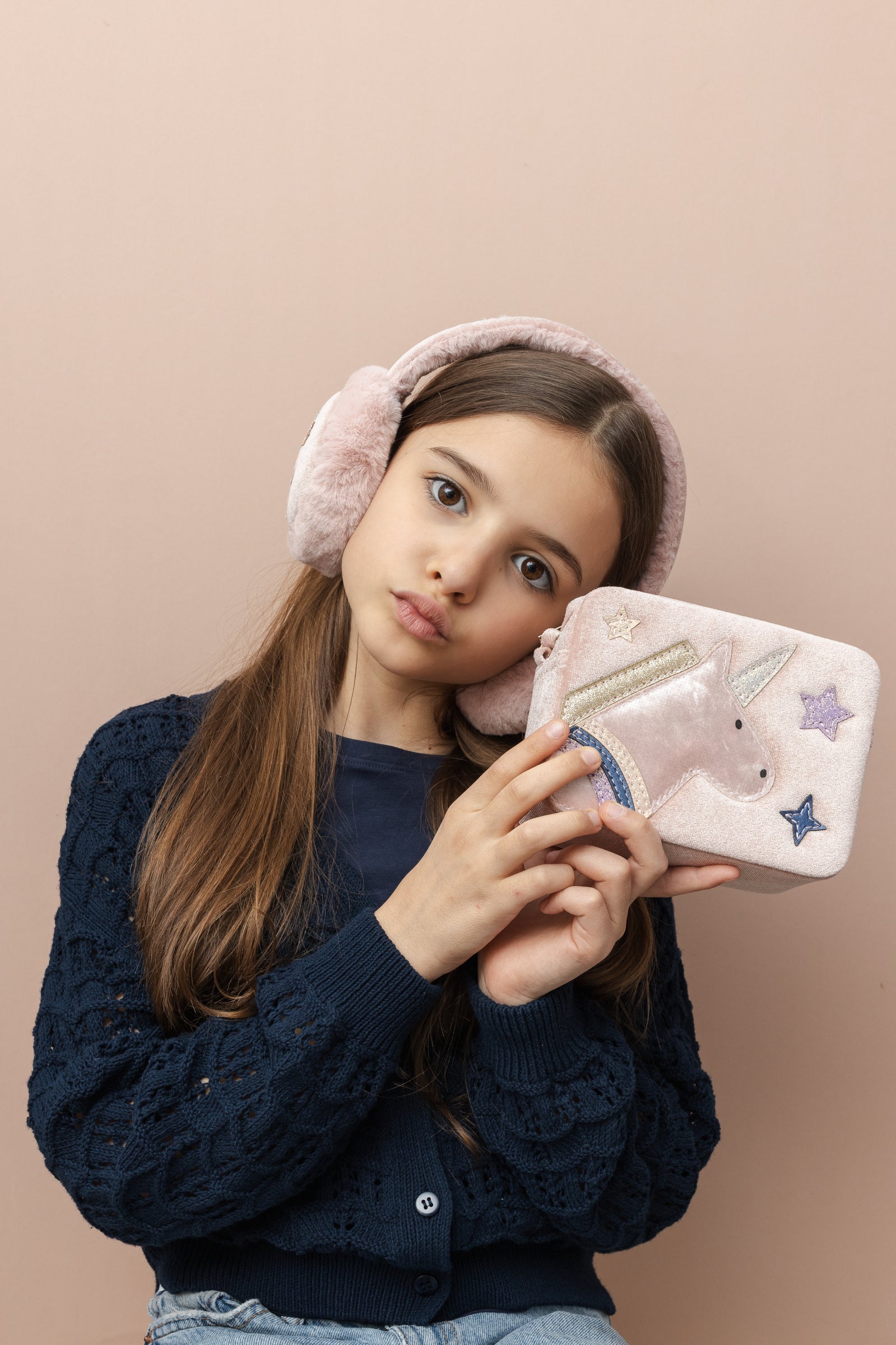 Young girl holding a unicorn-themed handbag with pink earmuffs on a beige background