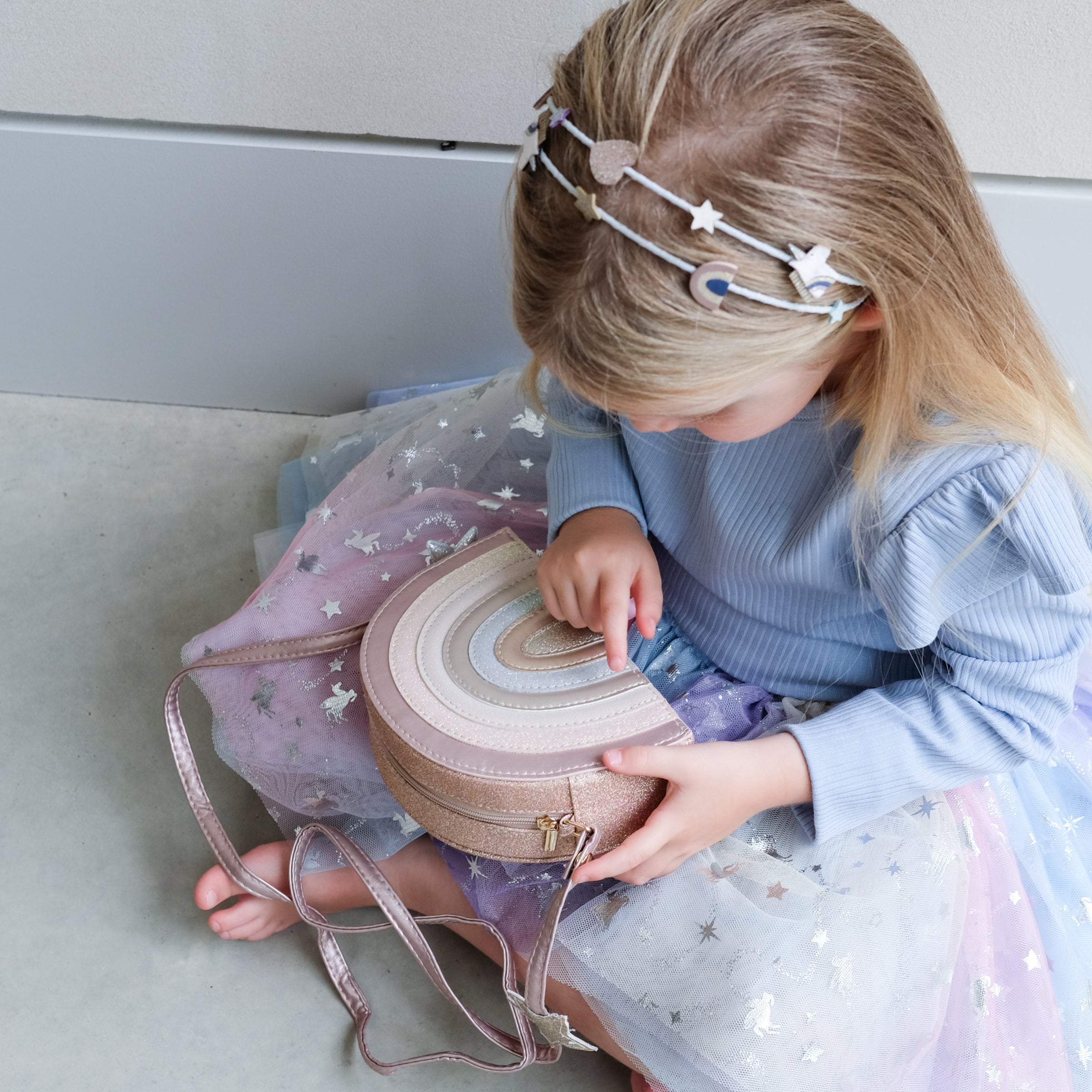 Young girl in a blue dress with a star headband, sitting on a light-colored surface, holding a pink rainbow shaped bag.